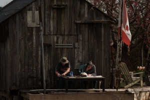 A couple sits on the deck in the afternoon spring sunshine. The gentleman is sharpening his axes, while his wife is enjoys a snack and reads the Sunday newspaper.